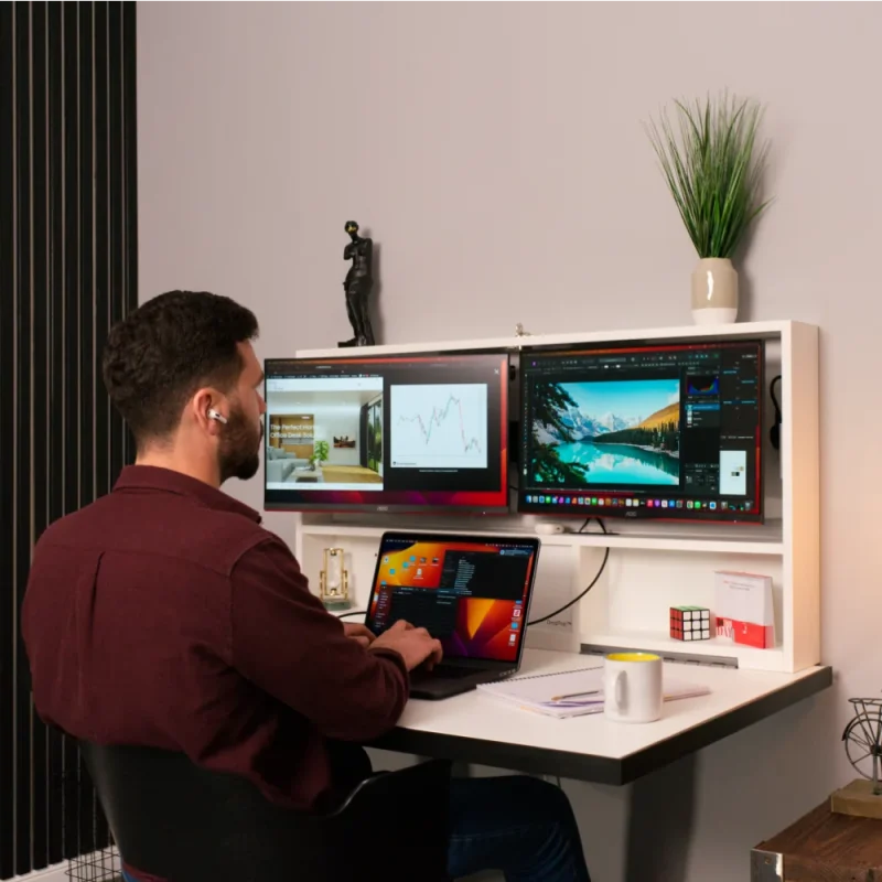 Man working at space saving fold out desk. Fitted with two screens and a shelf on top.
