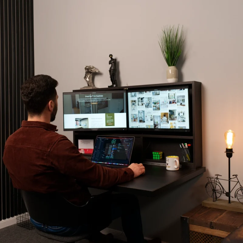 a man in a brown overskirt working on a black brown sorano droptop with dual screens with a MacBook Pro in a warm modern room