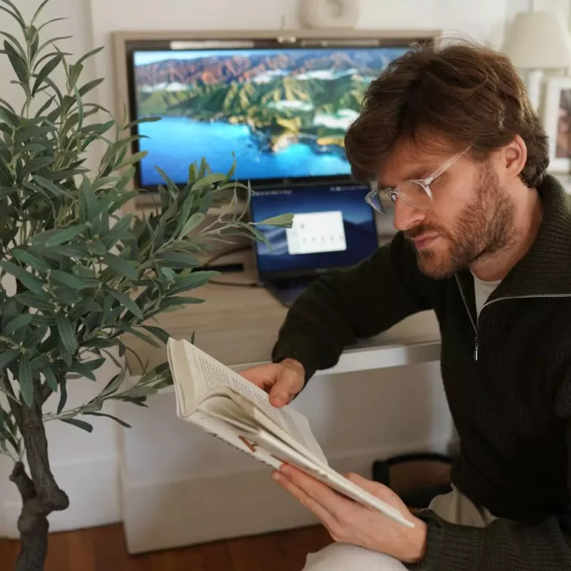 White male reading book in front of a wall desk with a laptop and monitor