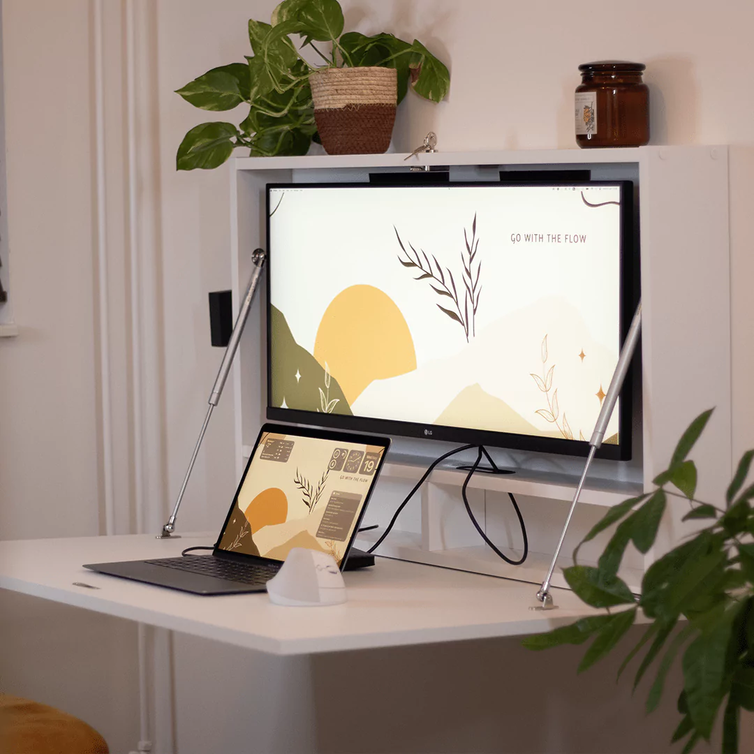 Wall-mounted fold-down desk with a large monitor and a laptop displaying a calming desktop background with the words ‘Go With The Flow.’ The workspace is decorated with plants and a candle, creating a serene and organised environment for work.