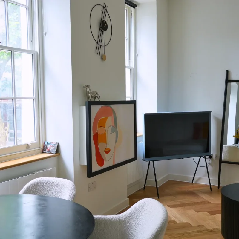 Minimalist living space featuring a wall-mounted artwork of an abstract face that doubles as a fold-down Murphy desk. The room is decorated with a modern clock, a TV on a stand, and soft chairs by the window, creating a functional yet stylish corner.