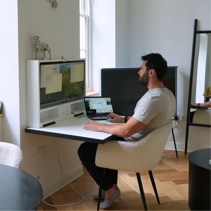 Person working at a wall-mounted fold-down desk with a large monitor and a laptop, seated in a cosy armchair. The modern workspace features a decorative sculpture, a mirror, and natural light from a nearby window, creating a stylish and functional home office setup.