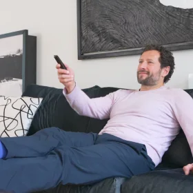 Person lounging on a sofa, casually holding a remote control, enjoying a moment of relaxation in a cosy living room. The setting highlights a comfortable work-life balance in a home environment.