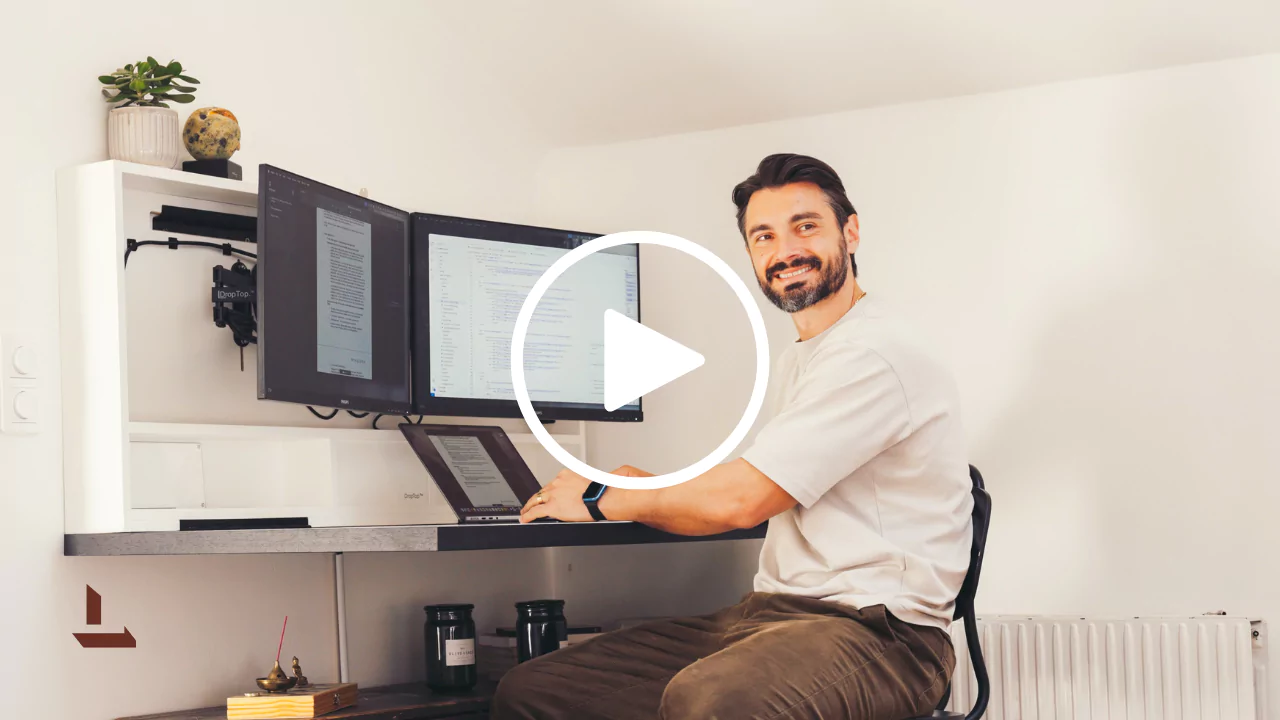Professional man working at a dual-monitor home office setup, smiling at the camera while typing. Modern ergonomic desk with two large screens showing code and documents, a laptop on a stand, and neatly arranged workspace accessories. Minimalist white wall background with indoor plant, globe, and candles, conveying productivity and clean design.