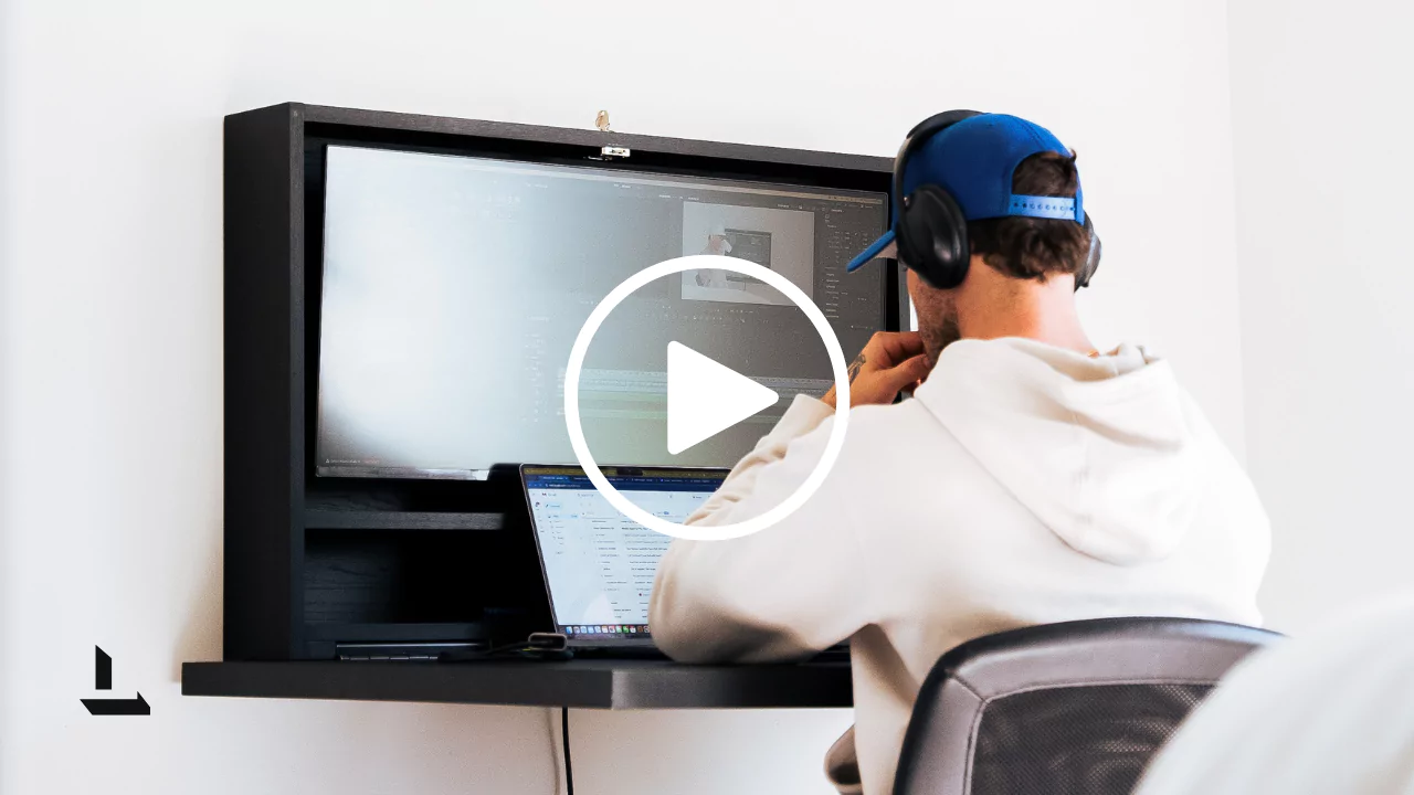 Creative video editor wearing a blue baseball cap and over-ear headphones, working at a minimalist black desk with a large wall-mounted monitor and laptop. Editing software is visible on the screen, showing a project timeline and preview window. The workspace is clean and organised, set against a plain white wall, conveying focus and professional content creation.
