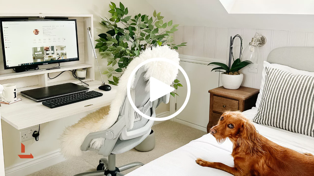 Cozy home office setup in a bright bedroom with a wall-mounted desk and monitor displaying a social media profile, a laptop, keyboard, and coffee mug on a light wood desk. Ergonomic grey chair draped with a white faux fur throw faces the workspace, while a golden-brown dog lies on the neatly made bed nearby. The room features green plants, a wooden nightstand with a potted orchid, striped cushions, and soft natural lighting, creating a warm and inviting remote work environment.